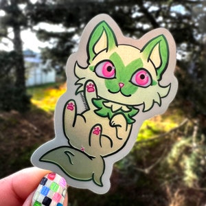 May include: A holographic sticker of a cartoon cat with green fur, pink paws, and pink eyes. The cat is smiling and has a green leaf around its neck.