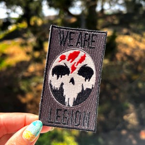 May include: A gray embroidered patch with a white skull and red splatter design. The text "WE ARE LEGION" is embroidered on the patch.