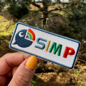 May include: White embroidered patch with the word "SIMP" in rainbow colors. The patch features a blue cartoon character with a rainbow behind it.