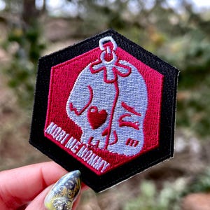 May include: A pink and white embroidered patch with a skull wearing a heart-shaped necklace. The patch is shaped like a hexagon and has black stitching around the edges. The text "MORTI ME MOMMY" is written in white on the patch.