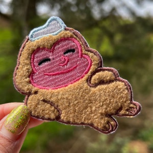 May include: A brown and pink embroidered patch of a smiling monkey wearing a blue and white hat. The monkey is lying on its back with its arms and legs outstretched.