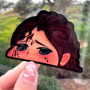 May include: A sticker of a cartoon character with brown hair, large eyes, and a red and white design. The character has blood dripping down their face.