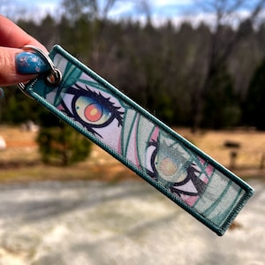 May include: A teal green keychain with a printed image of a pair of anime eyes with pink and yellow accents.