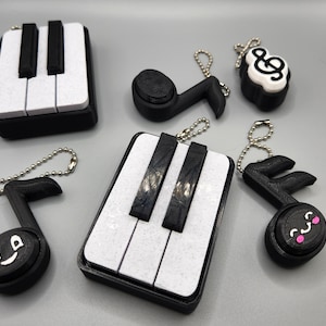May include: A collection of black and white piano key and musical note keychains. The keychains include a miniature piano keyboard and musical notes with smiling faces and treble clef designs. Each keychain has a silver chain.