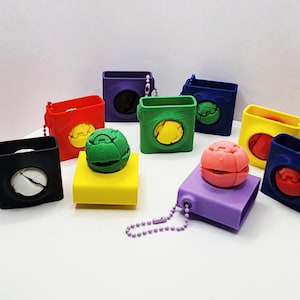 May include: A collection of colorful, square-shaped cases with matching spherical objects. The cases are in various colors, including black, red, green, yellow, blue, and purple. Each case has a circular opening and a small chain. The spherical objects are also in various colors.