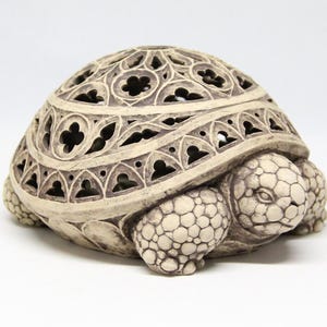 Puede incluir: Figura de tortuga de cerámica beige con un caparazón calado decorativo. El caparazón presenta intrincados patrones recortados, y la cabeza y las patas de la tortuga tienen un aspecto texturizado y escamoso. La figura está sobre un fondo blanco.