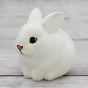 May include: A white ceramic rabbit figurine with black eyes and a pink nose. The rabbit is sitting on a white wooden surface.