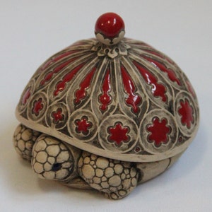 Puede incluir: Una caja de cerámica para joyas con forma de tortuga con una tapa roja. La tapa tiene un diseño decorativo y una pequeña perilla roja en la parte superior.