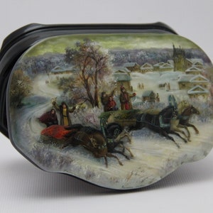 May include: A black lacquer box featuring a detailed hand-painted winter scene on its lid. The artwork depicts a horse-drawn carriage travelling through a snowy village landscape with trees, figures in traditional clothing, and a church in the background.