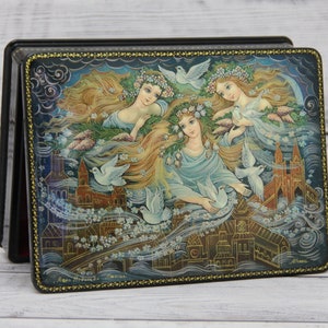 May include: A wooden box with a hinged lid, decorated with a colorful painting of three women with flowing hair and doves, set against a backdrop of a city skyline. The box is adorned with a gold trim.