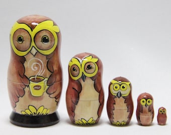 May include: Set of five brown and yellow painted wooden nesting dolls, each depicting an owl with a yellow beak and yellow eyes. The largest doll is holding a yellow cup with a brown swirl of steam coming from it.