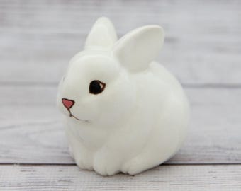 Puede incluir: Una figurita de conejo de cerámica blanca con ojos negros y nariz rosa. El conejo está sentado sobre una superficie de madera blanca.