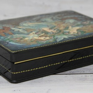 May include: A black wooden box with a hinged lid. The lid is decorated with a colorful painting of two women in a forest setting. The box has a gold trim around the edges.