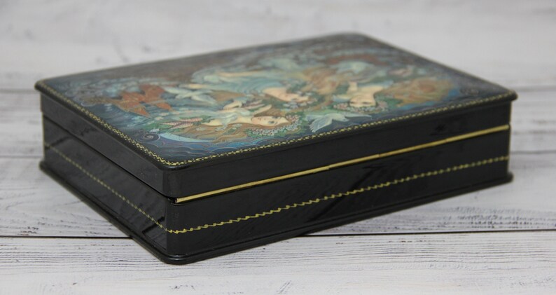 May include: A black wooden box with a hinged lid. The lid is decorated with a colorful painting of two women in a forest setting. The box has a gold trim around the edges.