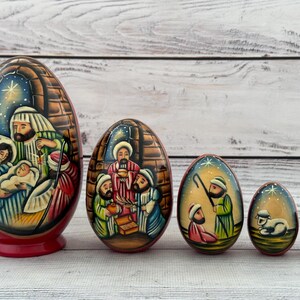 Peut inclure: Ensemble de cinq œufs de Pâques en bois peints à la main, représentant des scènes de la Nativité. Le plus grand œuf montre Marie, Joseph et l'enfant Jésus. Les œufs sont peints de couleurs vives sur un fond en bois clair.