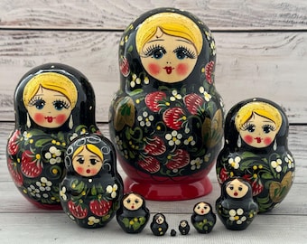 May include: A set of traditional Russian nesting dolls, or Matryoshka dolls, in various sizes. The dolls are painted black with floral and strawberry designs, and each doll features a painted face with blonde hair.