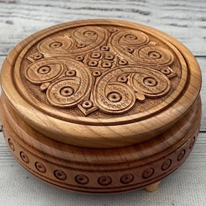 May include: A round wooden box with a carved lid featuring an intricate Celtic knot design. The box is light brown and has a decorative band of circles around the base. The box is approximately 4 inches in diameter.