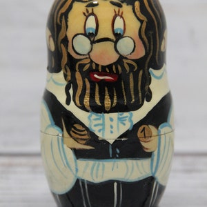 May include: A hand-painted wooden nesting doll, or matryoshka, depicting a man with a long black beard and glasses. He is wearing a black suit with a white shirt and a blue tie. The doll is numbered "1" on the bottom.