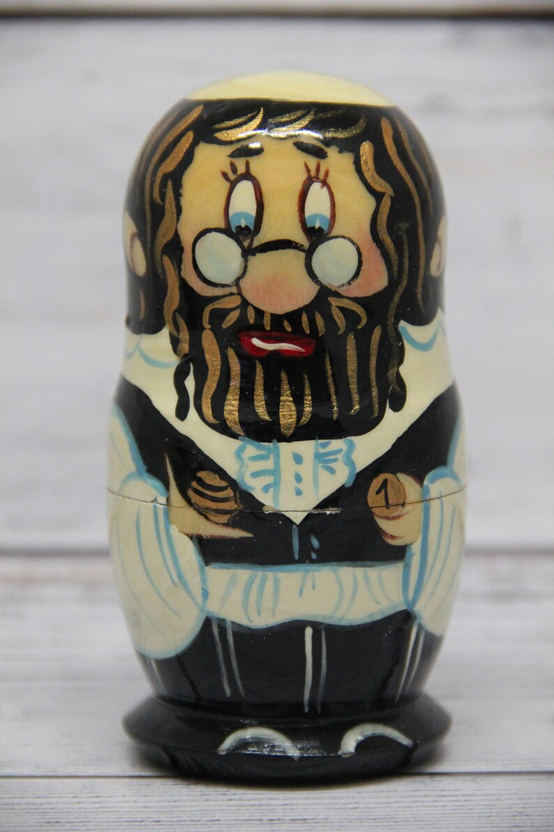 May include: A hand-painted wooden nesting doll, or matryoshka, depicting a man with a long black beard and glasses. He is wearing a black suit with a white shirt and a blue tie. The doll is numbered "1" on the bottom.