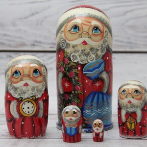 May include: Set of five traditional Russian nesting dolls, or matryoshka dolls, painted with Santa Claus designs. The largest doll is holding a blue bag, the second largest is holding a clock, the third largest is holding a blue bag, the fourth largest is holding a red bag, and the smallest is holding a red bag.