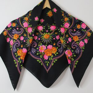 May include: A black square scarf with a floral pattern in pink, orange, and yellow. The scarf has a paisley design and is trimmed with a black border.