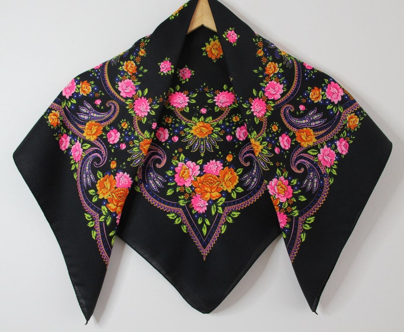 May include: A black square scarf with a floral pattern in pink, orange, and yellow. The scarf has a paisley design and is trimmed with a black border.