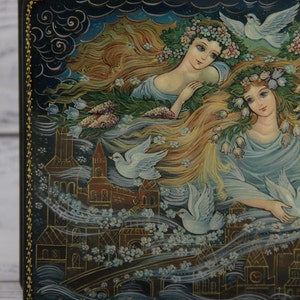 May include: A hand-painted wooden box with a dark blue background featuring two women with long flowing hair, wearing floral crowns and surrounded by white doves. The box is decorated with a gold border and a signature at the bottom.