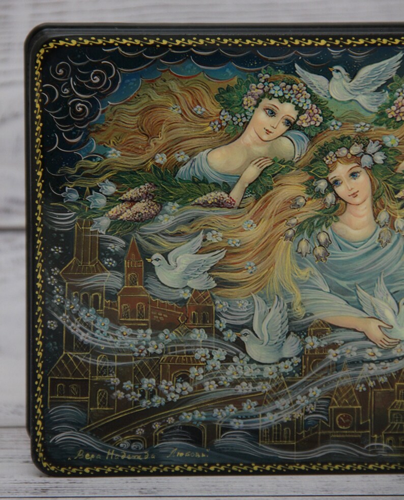 May include: A hand-painted wooden box with a dark blue background featuring two women with long flowing hair, wearing floral crowns and surrounded by white doves. The box is decorated with a gold border and a signature at the bottom.