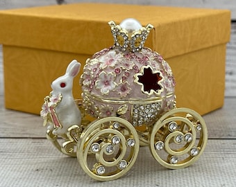May include: A decorative trinket box shaped like a pumpkin carriage with a white rabbit figure attached to the front. The carriage is light pink with gold trim, floral accents, and clear crystals. It features gold wheels with clear crystals.
