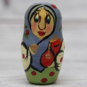 May include: A hand-painted wooden nesting doll, or matryoshka doll, with a blue and green dress, red accents, and a smiling face. The doll is holding a red object in its hand.