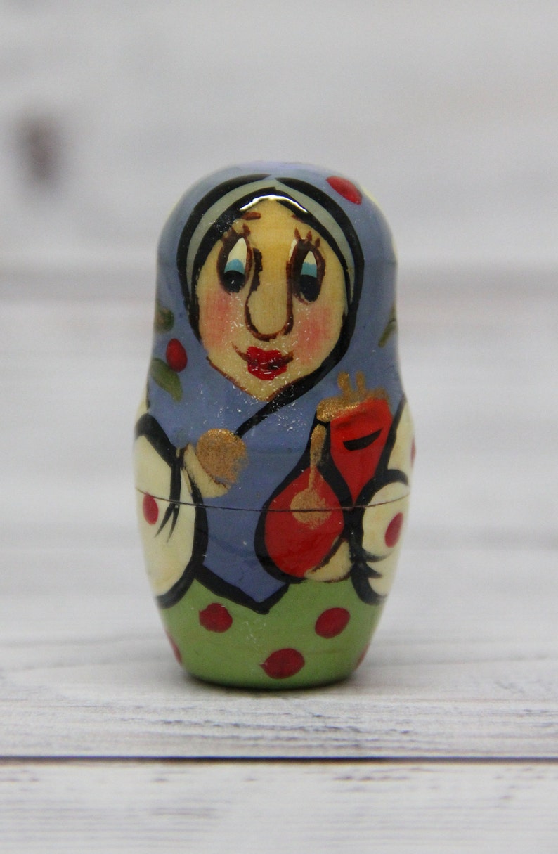 May include: A hand-painted wooden nesting doll, or matryoshka doll, with a blue and green dress, red accents, and a smiling face. The doll is holding a red object in its hand.