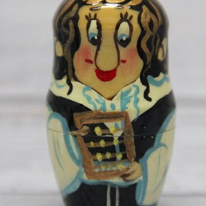 May include: A hand-painted wooden nesting doll, or matryoshka, depicting a woman with brown hair and a blue and white dress holding an abacus. The doll is painted in a whimsical style with a bright yellow background.