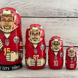 May include: Set of five red, white, and gold Kansas City Chiefs nesting dolls. Each doll features a different player wearing a helmet and jersey with their number. The largest doll has the number 15, the next largest has the number 87, the next has the number 10, the next has the number 32, and the smallest doll has the number 55.