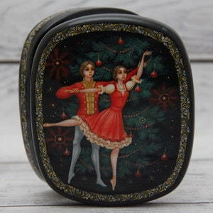 Op de afbeelding: Een zwarte gelakte houten doos met een geschilderd tafereel van een dansend stel voor een kerstboom. Het stel is gekleed in rood en wit, en de boom is versierd met ornamenten. De doos is versierd met een gouden rand.