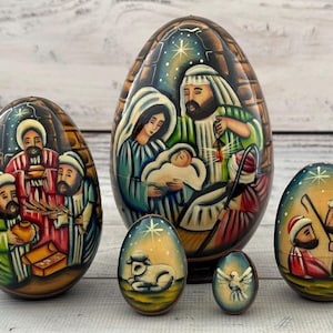 Peut inclure: Ensemble de six ornements en bois en forme d'œuf, chacun peint à la main avec des scènes religieuses. Le plus grand œuf représente la Nativité, tandis que d'autres montrent les Rois Mages et un agneau. La palette de couleurs comprend des bleus, des verts et des bruns.