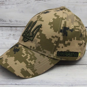 Puede incluir: Una gorra de béisbol de camuflaje con un tridente ucraniano bordado en verde y la palabra "UKRAINE" en letras amarillas.
