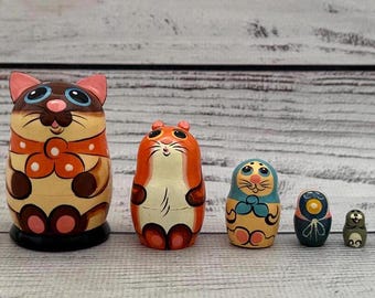 Könnte beinhalten: Ein Satz von fünf Holz-Matrjoschka-Puppen, jede mit einem anderen Tierdesign bemalt. Die größte Puppe ist eine Katze, gefolgt von einem Hamster, einer Katze, einer Eule und einem kleinen grünen Tier. Die Puppen sind in leuchtenden Farben bemalt.