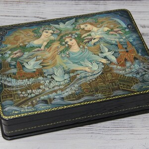 May include: A black wooden box with a hinged lid. The lid is decorated with a hand-painted scene of three women with flowing hair and white doves flying around them. The background is a blue sky with white clouds and a cityscape.