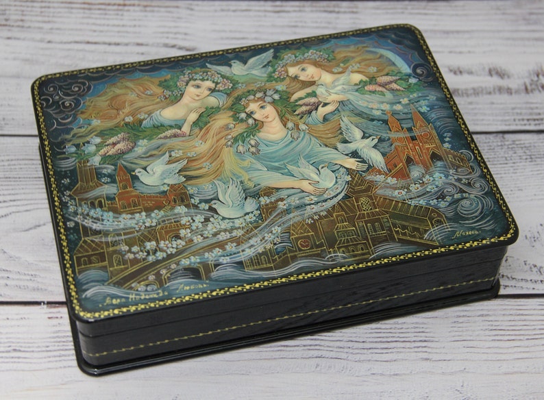 May include: A black wooden box with a hinged lid. The lid is decorated with a hand-painted scene of three women with flowing hair and white doves flying around them. The background is a blue sky with white clouds and a cityscape.