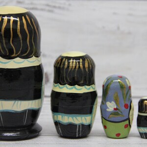 May include: A set of five traditional Russian nesting dolls, or matryoshka dolls. The largest doll is black with gold accents and a white apron. The other dolls are smaller and feature different colorful designs.
