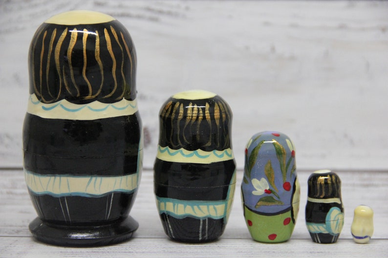 May include: A set of five traditional Russian nesting dolls, or matryoshka dolls. The largest doll is black with gold accents and a white apron. The other dolls are smaller and feature different colorful designs.