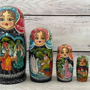 May include: A set of five traditional Russian nesting dolls, or matryoshka dolls. The largest doll is painted with a black background and features a scene of women in traditional clothing. The other dolls are painted with various scenes, including a swan, a castle, and a woman in a pink dress.