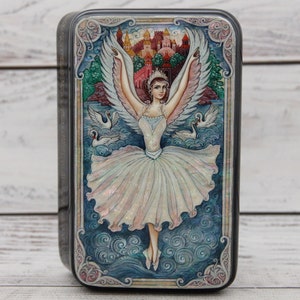 May include: A decorative metal box featuring a ballerina with angel wings, a crown, and a white tutu. The artwork includes swans, a castle, and a blue and green landscape. The box has a dark gray border.