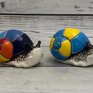 May include: Two ceramic snail figurines with glossy, multi-coloured shells. The left snail features a shell with segments of blue, red, orange, green, and yellow. The right snail has a shell with alternating segments of bright blue and yellow.