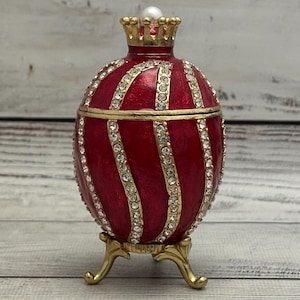Könnte beinhalten: Eine dekorative, rote eiförmige Schmuckschatulle mit goldenen Akzenten und klaren Kristallverzierungen. Das Ei hat eine goldene Krone und steht auf einem goldenen Ständer. Auf der Krone sitzt eine weiße Perle.