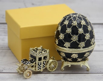 Peut inclure: Un œuf décoratif noir et or avec un motif floral, posé sur un socle doré. Une petite calèche dorée et une boîte cadeau jaune sont également dans l'image. L'œuf mesure environ 10 cm de haut.