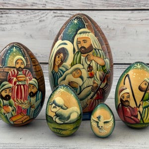 Peut inclure: Un ensemble de cinq œufs gigognes en bois peints à la main représentant une scène de nativité. Le plus grand œuf illustre la sainte famille, tandis que les autres montrent des bergers, un agneau et une colombe, ornés de couleurs vives.