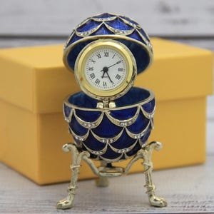 May include: A blue and gold jeweled egg clock with a white face and black hands. The egg is open and the clock is visible. The egg is sitting on a gold stand with three legs. The egg is in a yellow box.