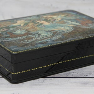 May include: A black wooden box with a hinged lid. The lid is decorated with a painted scene of three women in a forest setting. The box has a gold trim around the edges.