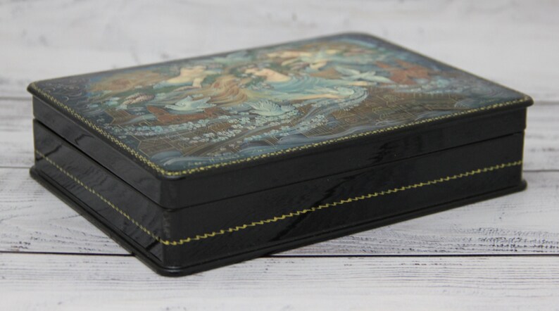 May include: A black wooden box with a hinged lid. The lid is decorated with a painted scene of three women in a forest setting. The box has a gold trim around the edges.